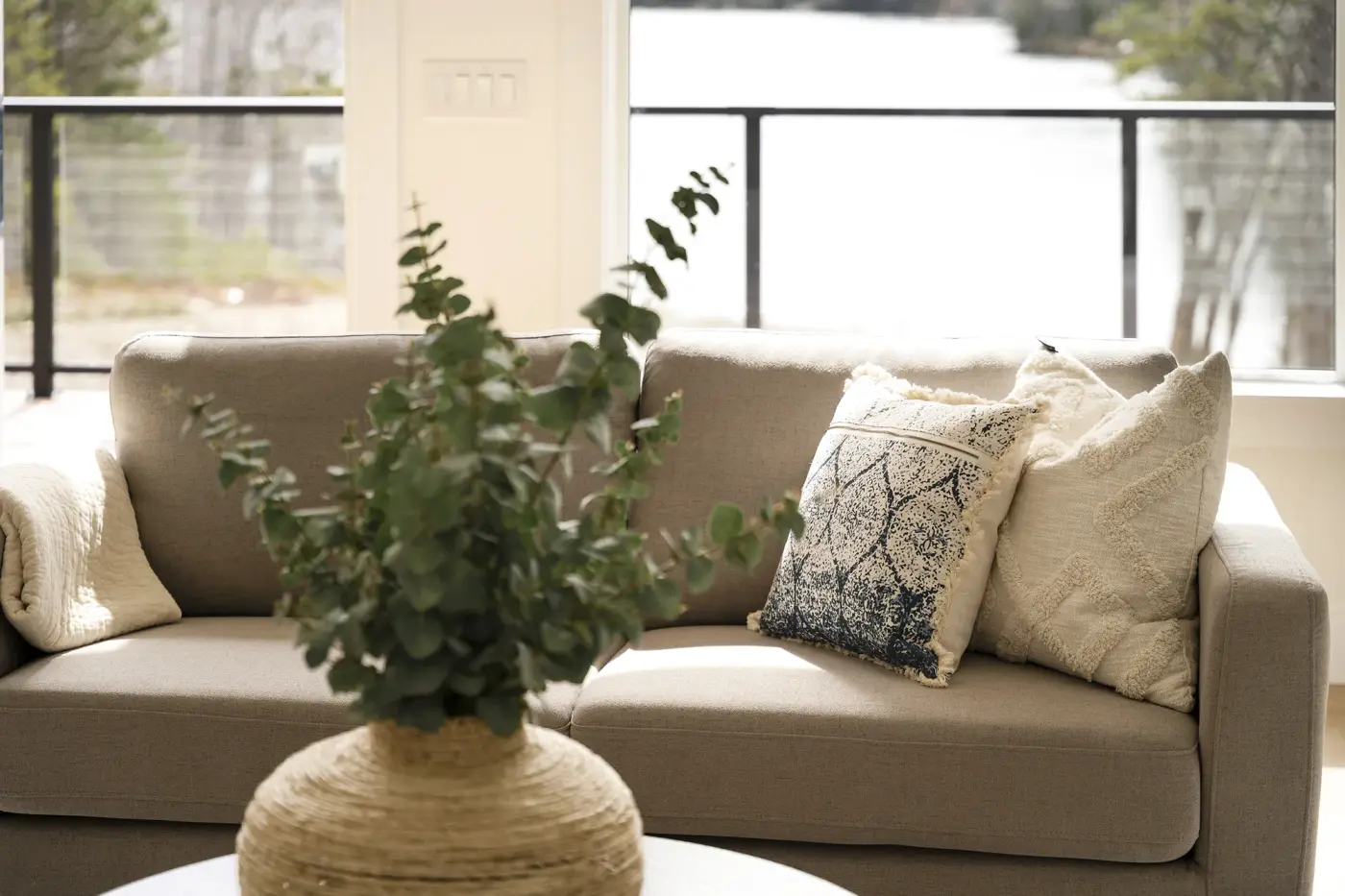 A linen sofa and eucalyptus by a wall of lake-facing windows in a Halifax home designed by MAD Design Interiors